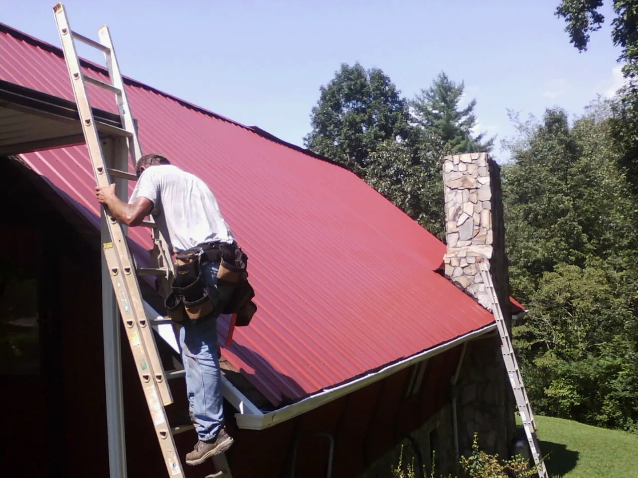 Roofer on a ladder beside a red metal roof and stone chimney — Soffit & Fascia Repair by Roofing Brothers in Seaton